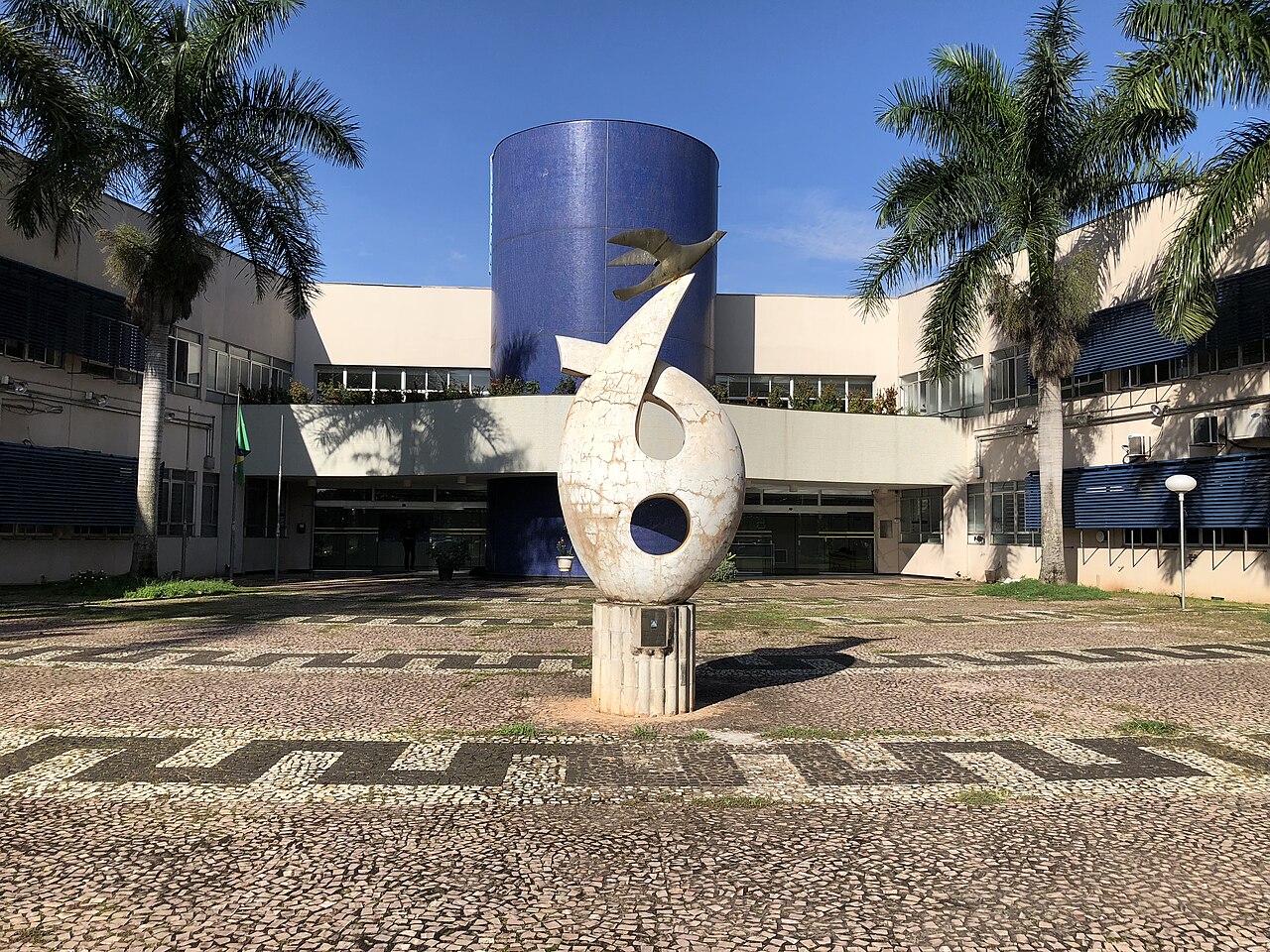 Fachada do prédio da reitoria da Universidade Federal de Goiás, com palmeiras e uma escultura no pátio.